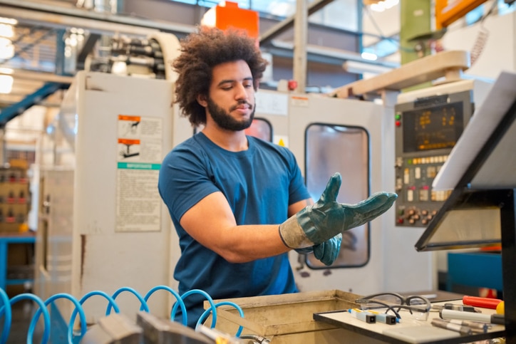 Junger Mann mit Bart und blauem Shirt zieht sich einen Arbeitshandschuh an und steht vor Maschinen in einer Industriehalle während seiner Zeitarbeit