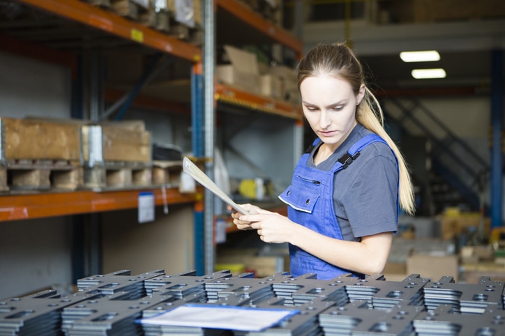 Junges blondes Mädchen beim Checken von Metall-Waren während ihres Zeitarbeit-Einsatzes im Lagerhaus einer Metallbaufirma