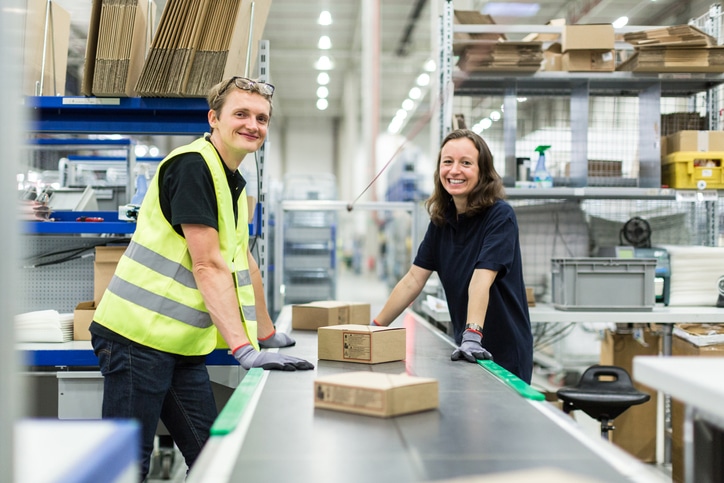 Ein junger Mann und eine junge Frauen stehen lächelnd am Fließband eines Paket-Lagers während ihres Zeitarbeits-Einsatzes.