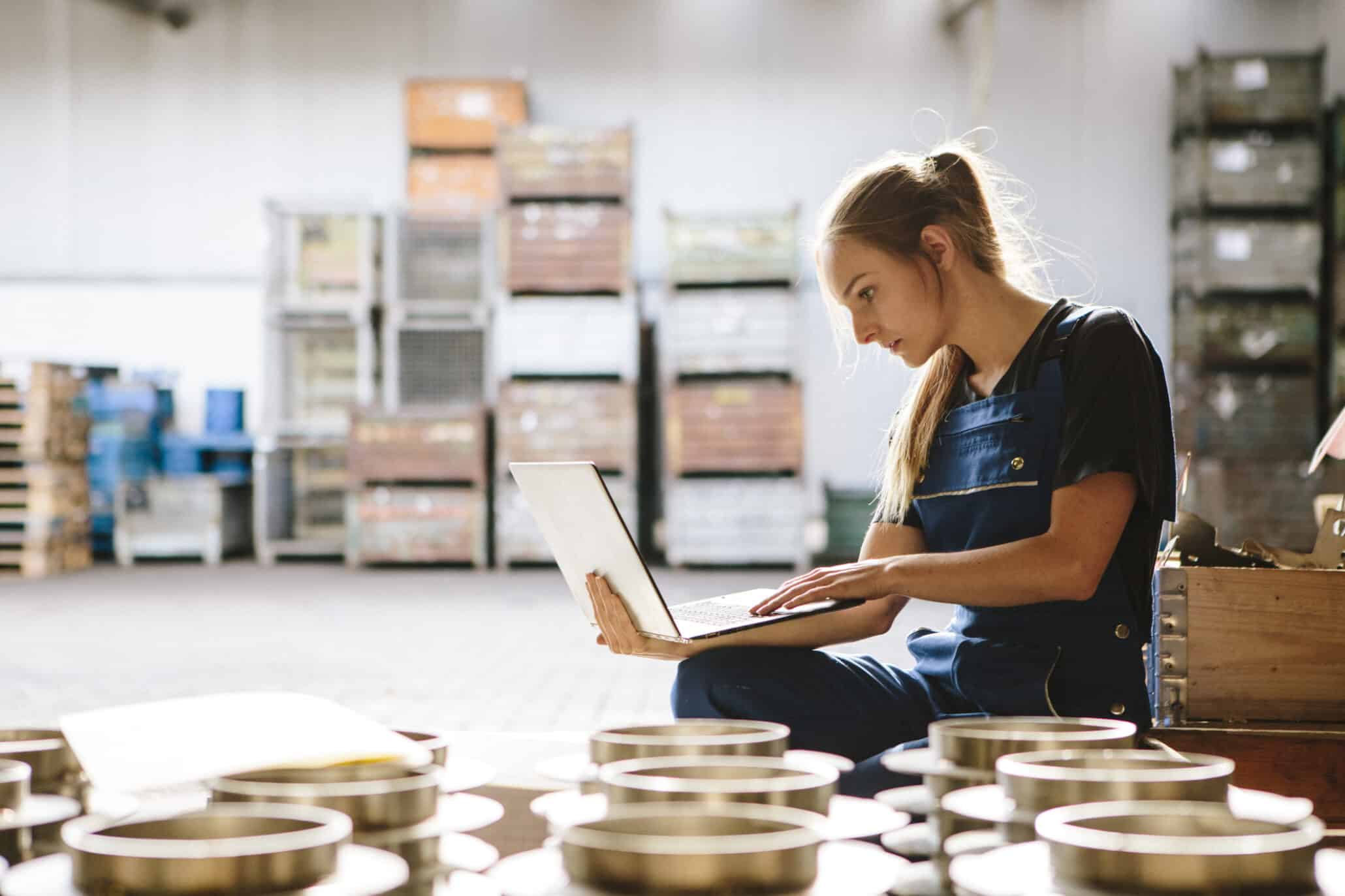 Junge Frau sitzt vor einem Laptop in einem Warenlager